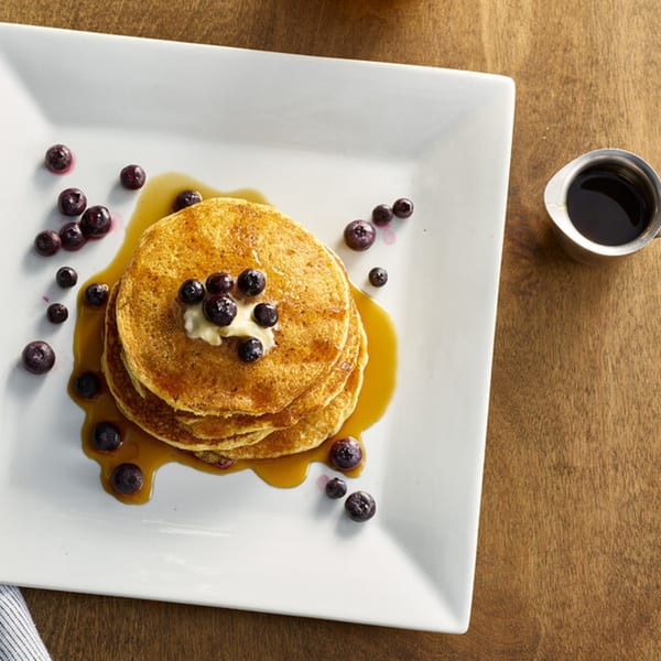 Stack of golden pancakes with butter, blueberries, and maple syrup on a white plate.