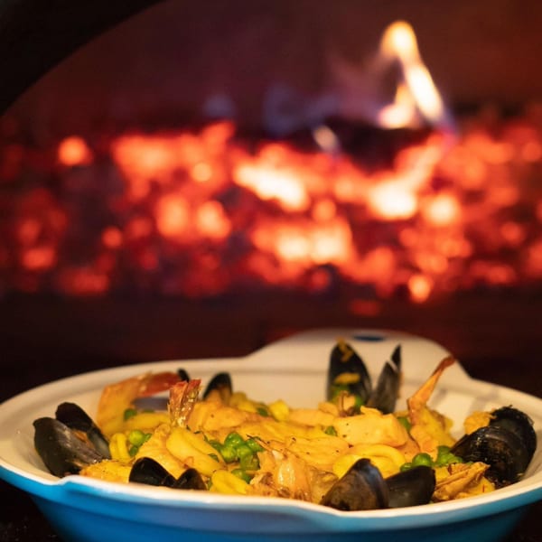 Seafood paella with mussels, shrimp, and peas in a white dish, with a fiery oven in the background.