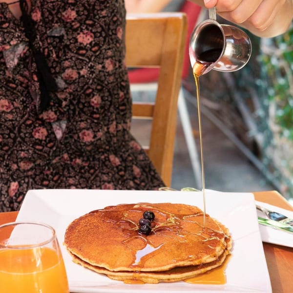 Maple syrup being poured over a stack of golden pancakes with blueberries and a glass of orange juice.