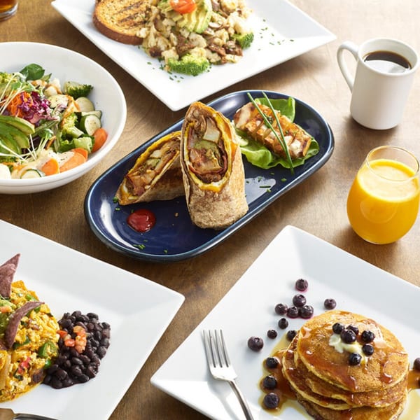 A table spread with various healthy dishes including pancakes, burritos, salad, and coffee.